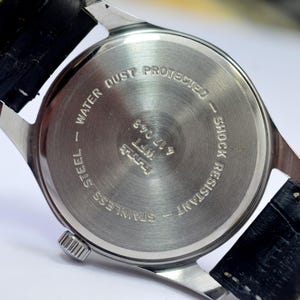May include: Close-up of the back of a stainless steel wristwatch with a black leather strap. The watch face is silver with the text "WATER DUST PROTECTED" and "SHOCK RESISTANT". The watch band is black leather.