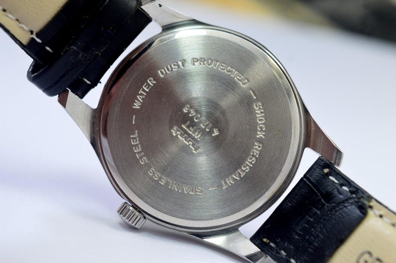 May include: Close-up of the back of a stainless steel wristwatch with a black leather strap. The watch face is silver with the text "WATER DUST PROTECTED" and "SHOCK RESISTANT". The watch band is black leather.