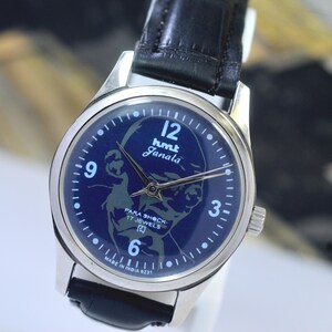 May include: A vintage wristwatch with a dark blue face featuring the text "hmt Janala" and an image of a person. The watch has silver hands and a silver case, with a black leather strap. The watch face also includes the text "PARA SHOCK 17 JEWELS".