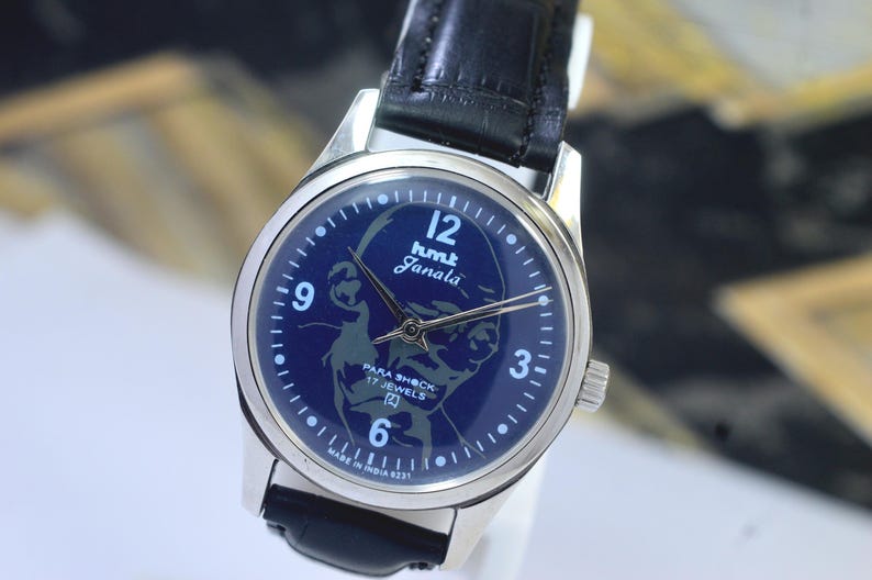 May include: A vintage wristwatch with a dark blue face featuring the text "hmt Janala" and an image of a person. The watch has silver hands and a silver case, with a black leather strap. The watch face also includes the text "PARA SHOCK 17 JEWELS".