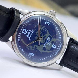 May include: A vintage wristwatch with a dark blue face featuring a stylized illustration and the text "hmt Janata". The watch has silver-toned hands and a silver case, paired with a black leather strap.