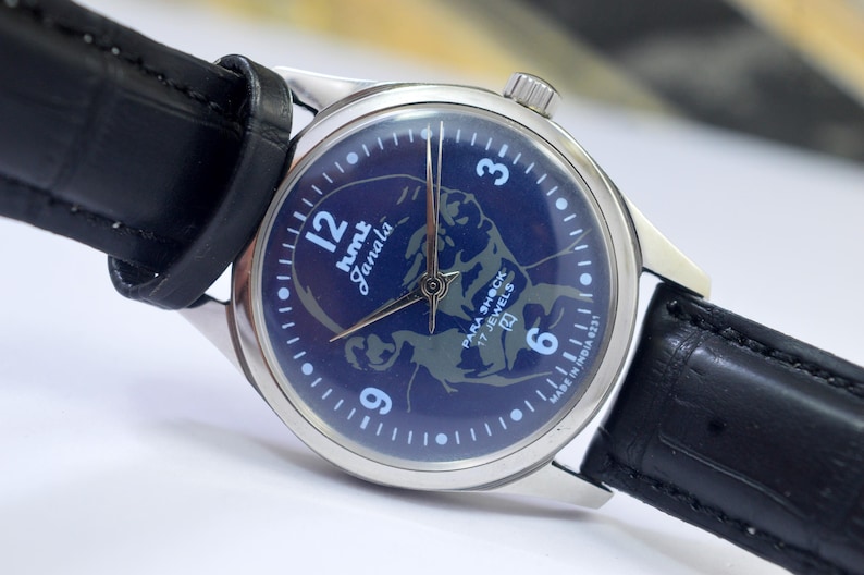 May include: A vintage wristwatch with a dark blue face featuring a stylized illustration and the text "hmt Janata". The watch has silver-toned hands and a silver case, paired with a black leather strap.