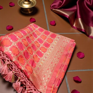 Peut inclure: Un textile à motifs rose et orange vif, avec des détails dorés et une bordure à franges. Le tissu est plié, révélant un motif complexe. Une bougie dans un support en laiton et des pétales de rose sont également visibles.