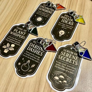 May include: A collection of decorative potion bottle pendants with various colors and text. Each pendant has a unique design and text, such as "Tonic of Plant Whispers" and "Serum of Serene Secrets". The pendants are on a wooden surface.