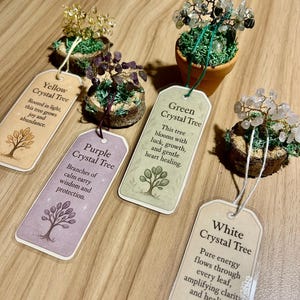 May include: A collection of crystal trees with tags. The trees feature wire branches adorned with crystals in yellow, purple, green, and white. Each tree sits on a small, moss-covered base or in a terracotta pot. The tags describe the tree's properties.