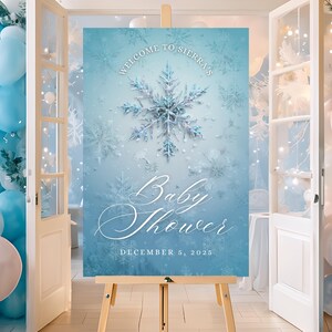 Może przedstawiać: Znak baby shower na sztaludze o tematyce zimowej. Znak jest jasnoniebieski z motywem płatka śniegu i napisami "Welcome to Sierra's", "Baby Shower" i "December 5, 2025". Balony w odcieniach niebieskiego i białego zdobią tło.