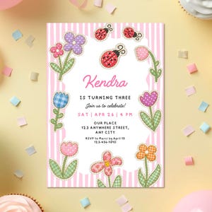 Könnte beinhalten: Eine Geburtstagseinladung mit rosa und weißen Streifen, die Blumen- und Marienkäfer-Designs zeigt. Die Einladung lautet "Kendra wird drei" mit Veranstaltungsdetails. Konfetti und Cupcakes sind ebenfalls zu sehen.