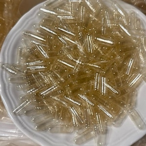 以下が含まれることがあります： 白いセラミック皿に、多数の透明な空のゼラチンカプセルが詰められています。カプセルは円筒形で丸みを帯びた端があり、密集して重なり合うパターンで配置されています。カプセルは半透明です。