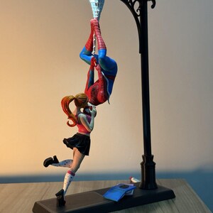 May include: A black lamp post with a light at the top, featuring a figurine of Spider-Man hanging upside down and a girl in a pink top and black skirt. The base is rectangular and black.