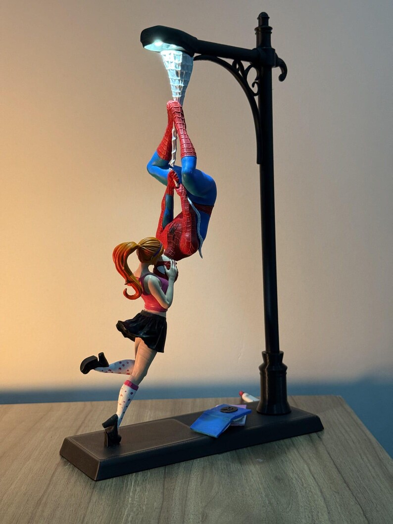 May include: A black lamp post with a light at the top, featuring a figurine of Spider-Man hanging upside down and a girl in a pink top and black skirt. The base is rectangular and black.