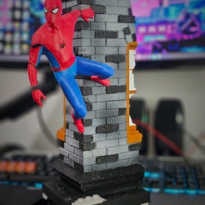 May include: A red, blue, and black Spider-Man figurine climbing a gray brick structure. The figurine is posed mid-climb, with one hand and foot gripping the brick. The base is black with white accents. The background is blurred.