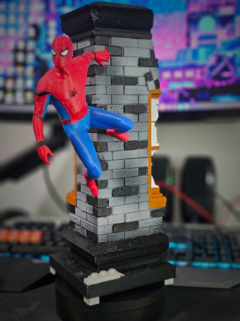 May include: A red, blue, and black Spider-Man figurine climbing a gray brick structure. The figurine is posed mid-climb, with one hand and foot gripping the brick. The base is black with white accents. The background is blurred.