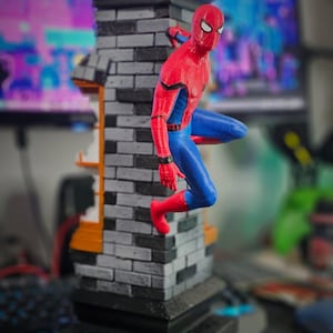 May include: A detailed figurine of Spider-Man in a dynamic pose, clinging to a brick structure. The figure is clad in a red and blue suit, with a black and gray brick tower base. The background is blurred.
