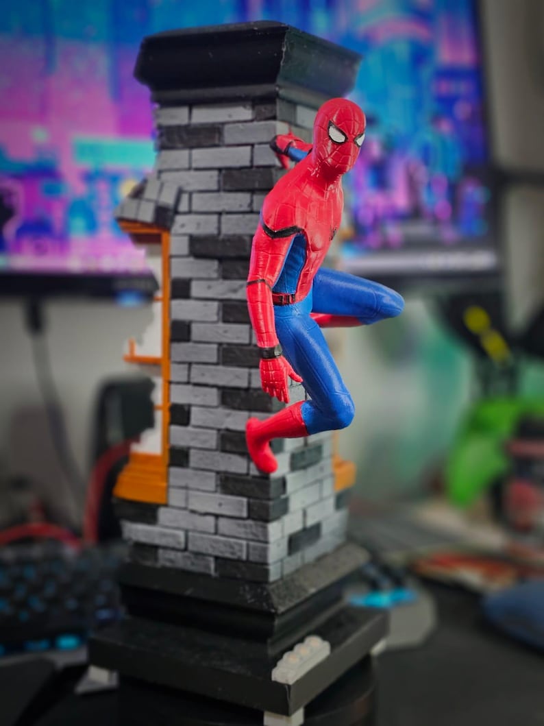May include: A detailed figurine of Spider-Man in a dynamic pose, clinging to a brick structure. The figure is clad in a red and blue suit, with a black and gray brick tower base. The background is blurred.