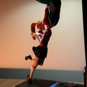 May include: A figurine of Spider-Man kissing a woman upside down. The woman wears a pink top, black skirt, and polka-dot socks. Books are on the base of the figurine.