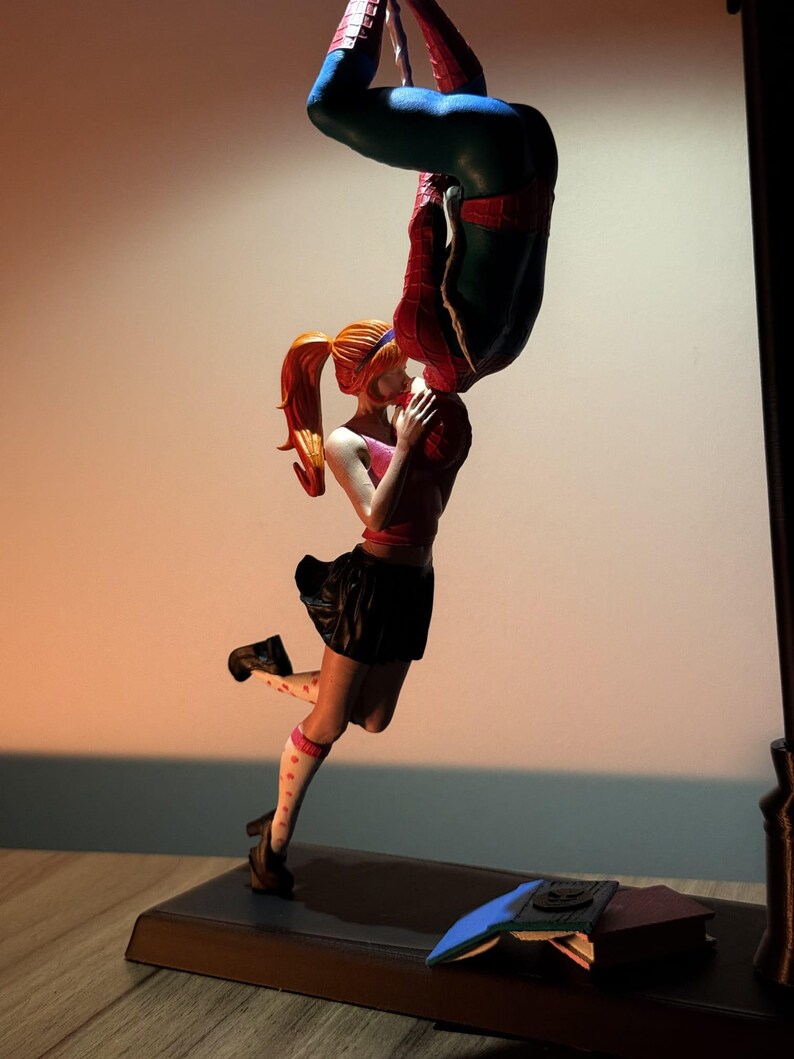 May include: A figurine of Spider-Man kissing a woman upside down. The woman wears a pink top, black skirt, and polka-dot socks. Books are on the base of the figurine.