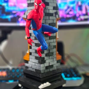 May include: A Spider-Man figurine in a dynamic pose, clinging to a brick-patterned structure. The figure is clad in a red and blue suit, with a black spider emblem on the chest. The base is black, and the overall design is detailed and vibrant.