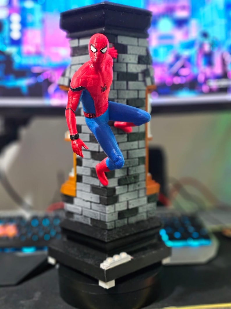 May include: A Spider-Man figurine in a dynamic pose, clinging to a brick-patterned structure. The figure is clad in a red and blue suit, with a black spider emblem on the chest. The base is black, and the overall design is detailed and vibrant.