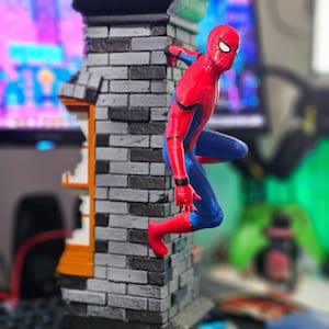 May include: A Spider-Man figurine in a dynamic pose, clinging to a brick building facade. The figure is in a classic red and blue suit. The building has a gray brick pattern with black and orange accents.