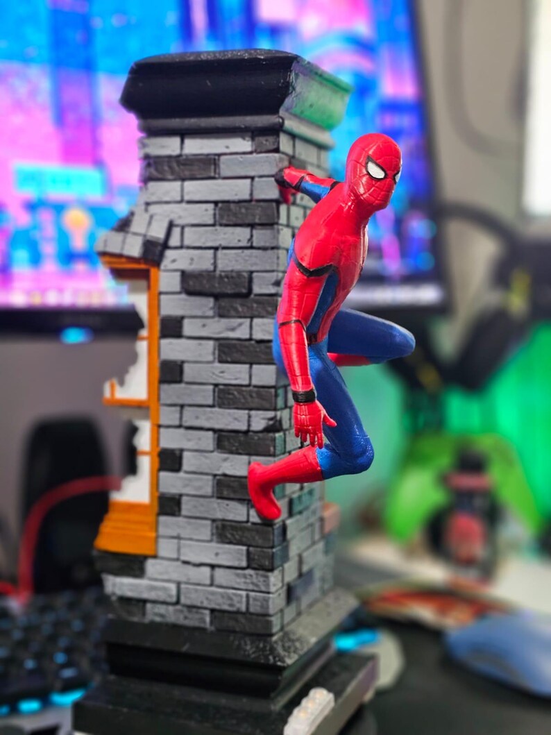 May include: A Spider-Man figurine in a dynamic pose, clinging to a brick building facade. The figure is in a classic red and blue suit. The building has a gray brick pattern with black and orange accents.