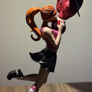 May include: Figurine of a woman in a pink top and black skirt kissing a figure in a Spider-Man suit. The woman has red hair in a ponytail and is wearing white socks with heart patterns. The Spider-Man figure is upside down.