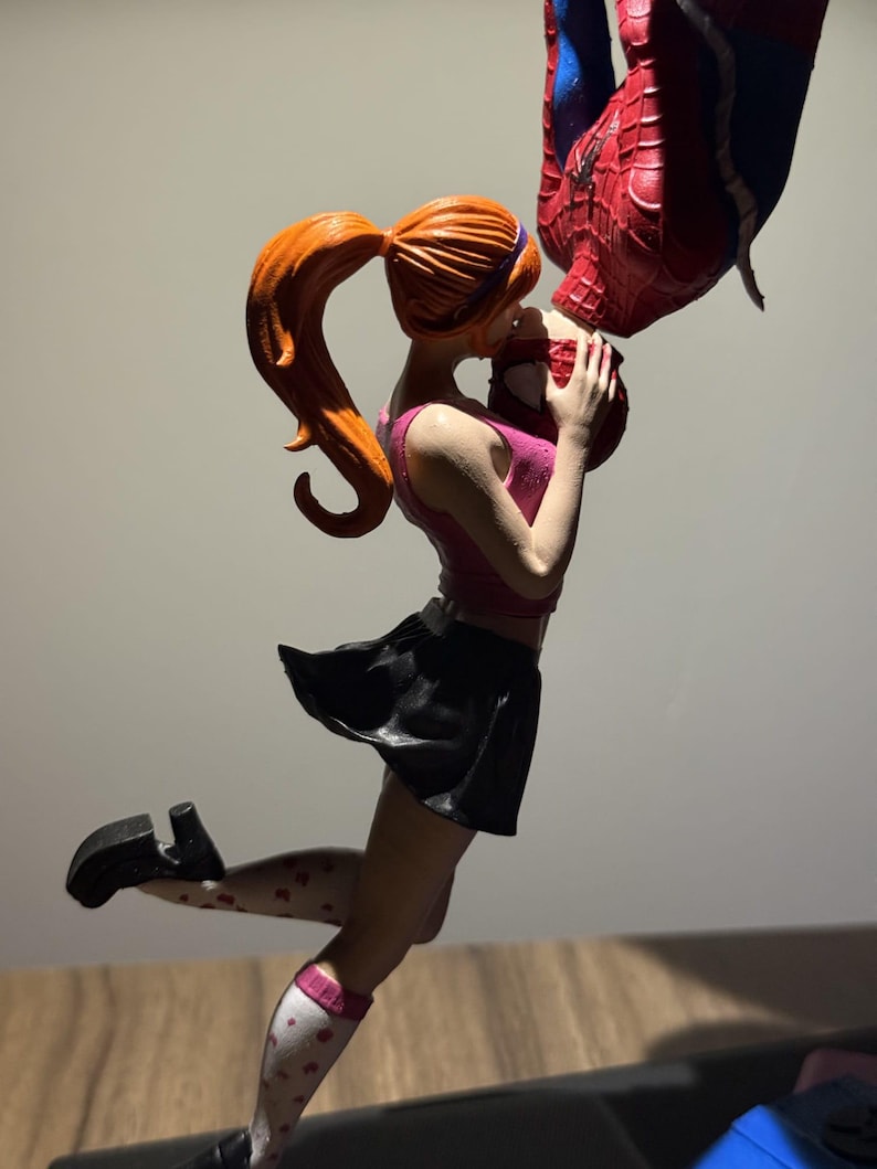 May include: Figurine of a woman in a pink top and black skirt kissing a figure in a Spider-Man suit. The woman has red hair in a ponytail and is wearing white socks with heart patterns. The Spider-Man figure is upside down.