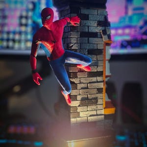 May include: A Spider-Man action figure in a dynamic pose, clinging to a brick wall. The figure is clad in a red and blue suit, with a black web design. The wall is gray and brown, with a black top. The scene is illuminated by a spotlight.
