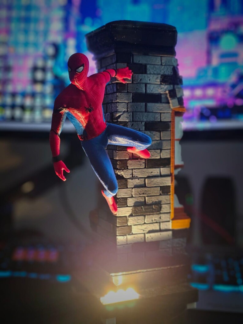 May include: A Spider-Man action figure in a dynamic pose, clinging to a brick wall. The figure is clad in a red and blue suit, with a black web design. The wall is gray and brown, with a black top. The scene is illuminated by a spotlight.