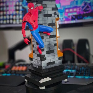 May include: A Spider-Man figurine in a dynamic pose, clinging to a brick-patterned structure. The figure is clad in a red and blue suit, with a black base. The structure has a black top and orange accents, creating a striking contrast.