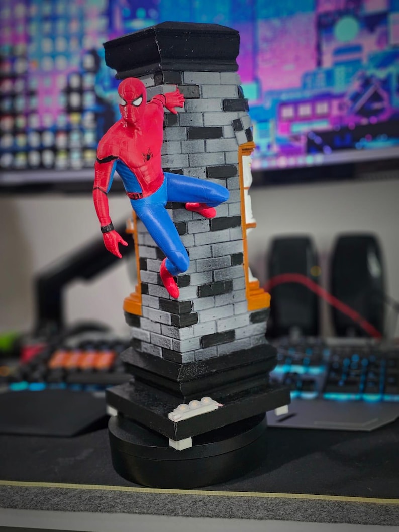 May include: A Spider-Man figurine in a dynamic pose, clinging to a brick-patterned structure. The figure is clad in a red and blue suit, with a black base. The structure has a black top and orange accents, creating a striking contrast.