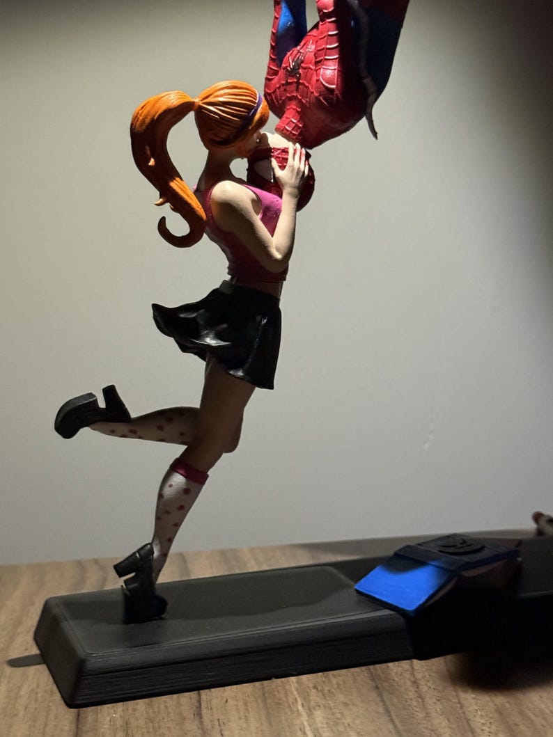 May include: Figurine of a scene from Spider-Man. A woman with red hair in a ponytail, wearing a pink top and black skirt, kisses Spider-Man, who is hanging upside down. The figurine is on a black base.