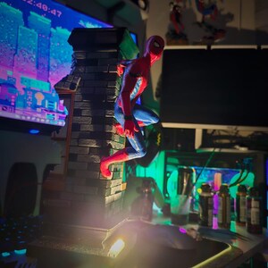 May include: A red and blue Spider-Man action figure perched on a brick structure. The figure is in a dynamic pose, with a cityscape visible in the background. The time 22:48:47 is displayed on a monitor.