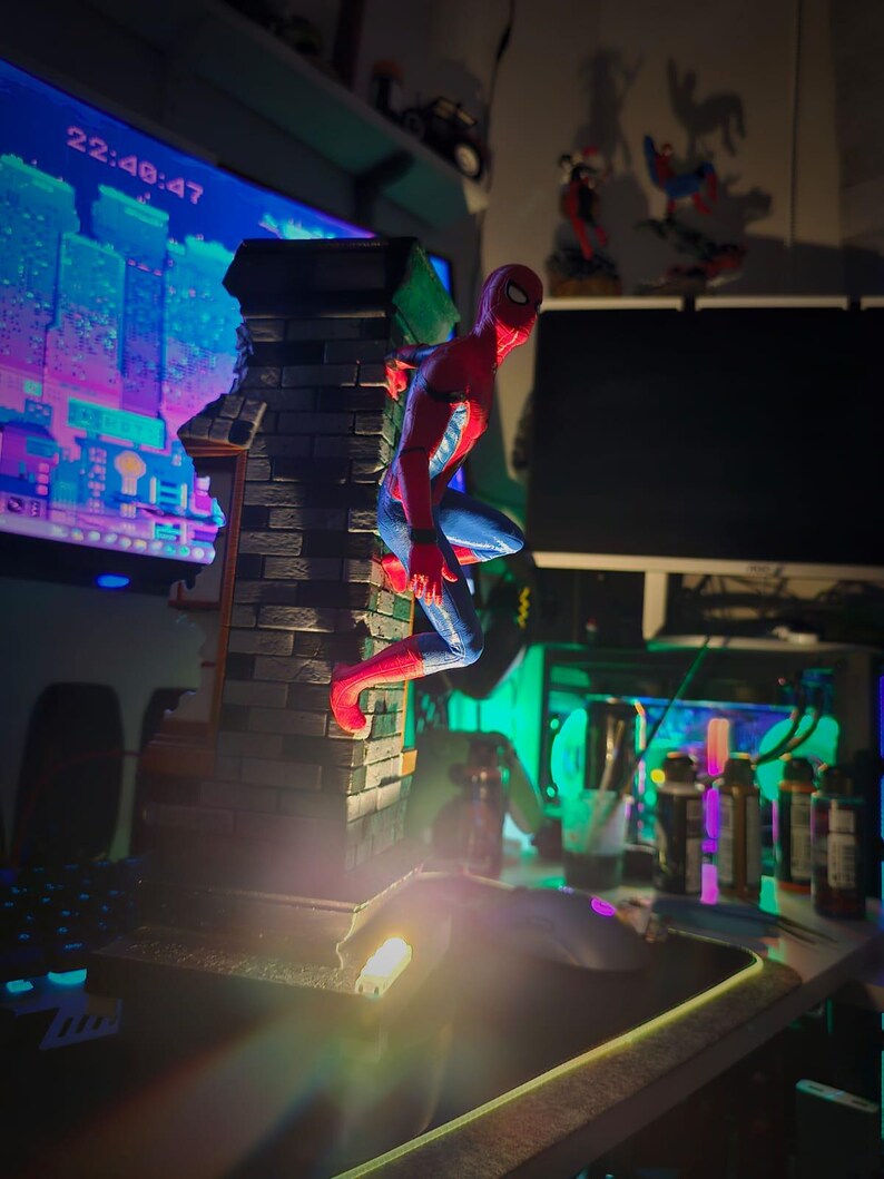 May include: A red and blue Spider-Man action figure perched on a brick structure. The figure is in a dynamic pose, with a cityscape visible in the background. The time 22:48:47 is displayed on a monitor.