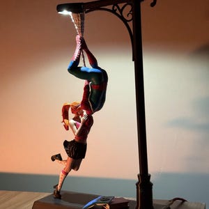 May include: A decorative lamp featuring a Spiderman figure hanging upside down, kissing a woman. The lamp has a street light design with a dark brown base and pole. The figures are in vibrant colours.