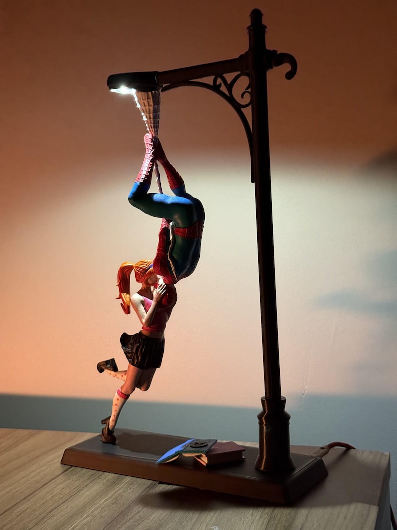 May include: A decorative lamp featuring a Spiderman figure hanging upside down, kissing a woman. The lamp has a street light design with a dark brown base and pole. The figures are in vibrant colours.