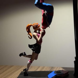 May include: A statue of Spider-Man kissing a woman upside down. The woman is in a pink top, black skirt, and white socks with red hearts. The statue is mounted on a black base.