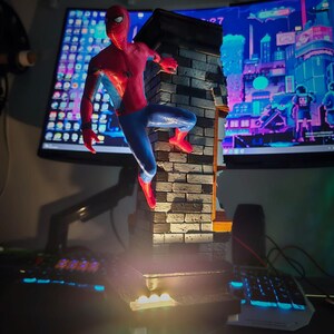 May include: A detailed figurine of Spider-Man in a dynamic pose, clinging to a brick wall. The figure is predominantly red and blue, with a black spider emblem. The wall is textured with realistic brickwork. The base has a light.