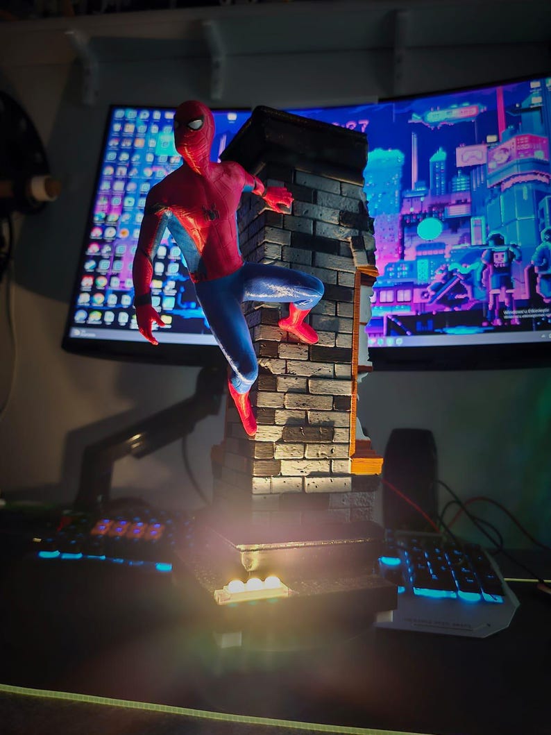 May include: A detailed figurine of Spider-Man in a dynamic pose, clinging to a brick wall. The figure is predominantly red and blue, with a black spider emblem. The wall is textured with realistic brickwork. The base has a light.