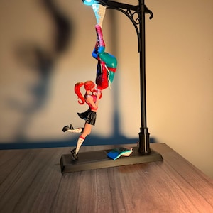May include: A black street lamp figurine featuring a Spiderman figure hanging upside down and a woman figure in a black skirt and red top. The lamp has a blue light. A small book is on the base.