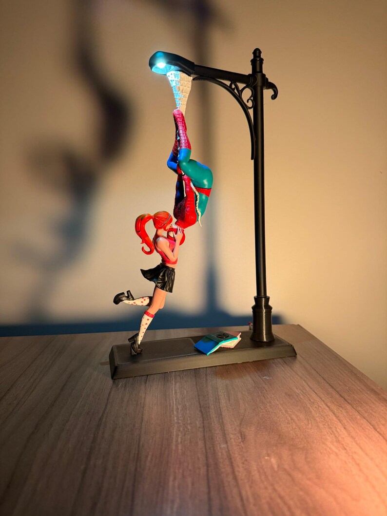 May include: A black street lamp figurine featuring a Spiderman figure hanging upside down and a woman figure in a black skirt and red top. The lamp has a blue light. A small book is on the base.