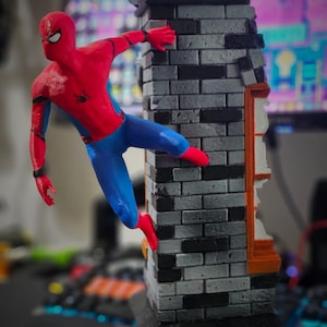 May include: A Spider-Man figurine in a dynamic pose, clinging to a brick wall. The figure is in a classic red and blue suit with black web detailing. The wall is gray and black, with an orange and white window frame.