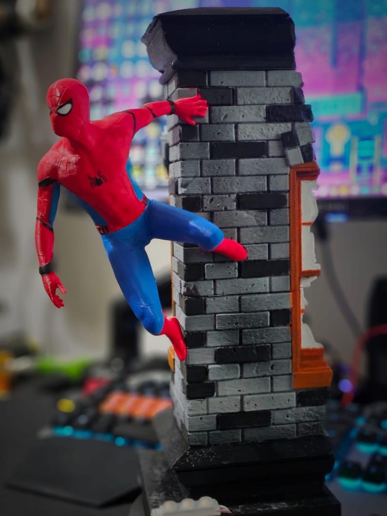 May include: A Spider-Man figurine in a dynamic pose, clinging to a brick wall. The figure is in a classic red and blue suit with black web detailing. The wall is gray and black, with an orange and white window frame.