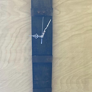 May include: A dark wood, vertical wall clock with silver numerals at the top and bottom. The clock has silver hands and metal bands. It is mounted on a light wood surface. The clock's design is rustic.