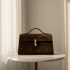 K&ouml;nnte beinhalten: Braune Wildlederhandtasche mit goldfarbenem Verschluss und Griff. Die rechteckige Tasche hat einen Klappverschluss und steht auf einem wei&szlig;en Marmortisch. Das Design der Tasche ist klassisch und elegant.