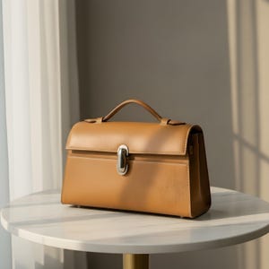 K&ouml;nnte beinhalten: Eine hellbraune Lederhandtasche mit einem oberen Griff und einem silberfarbenen Verschluss. Die strukturierte Tasche hat eine rechteckige Form mit einem Klappenverschluss und steht auf einem wei&szlig;en Marmortisch. Die Tasche ist neutralfarben.