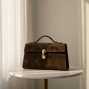 K&ouml;nnte beinhalten: Eine braune Handtasche mit goldenem Verschluss und Griff. Die rechteckige Tasche hat ein strukturiertes Design und steht auf einem wei&szlig;en Marmortisch. Der Griff ist dunkelbraun, und die Tasche scheint aus Wildleder oder einem &auml;hnlichen Material zu sein.