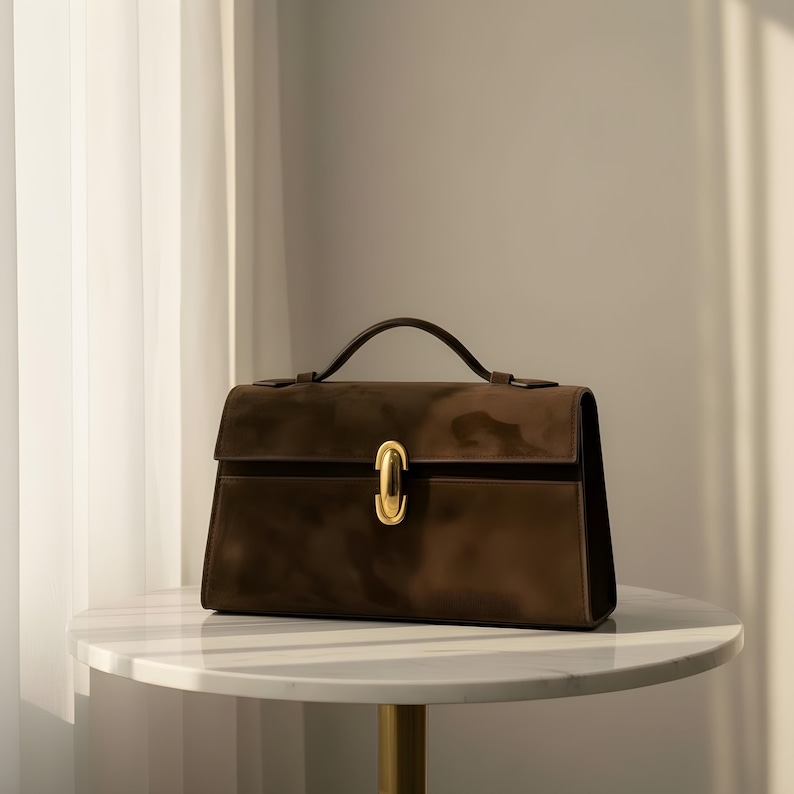 K&ouml;nnte beinhalten: Eine braune Handtasche mit goldenem Verschluss und Griff. Die rechteckige Tasche hat ein strukturiertes Design und steht auf einem wei&szlig;en Marmortisch. Der Griff ist dunkelbraun, und die Tasche scheint aus Wildleder oder einem &auml;hnlichen Material zu sein.