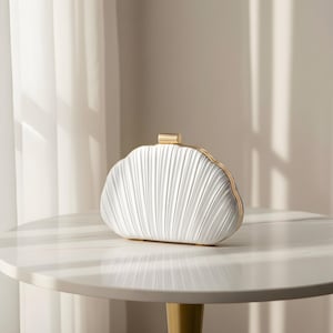 Peut inclure: Une pochette blanche avec un design plissé et une bordure dorée. La pochette est posée sur une table blanche avec un pied doré. L'arrière-plan est de couleur neutre avec un rideau blanc.