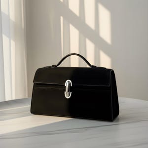 K&ouml;nnte beinhalten: Eine schwarze Handtasche mit einem oberen Griff und einem silberfarbenen Verschluss. Die rechteckige Tasche hat einen Klappenverschluss und steht auf einer wei&szlig;en Oberfl&auml;che. Die Tasche ist aus glattem Material gefertigt und hat ein strukturiertes Design.