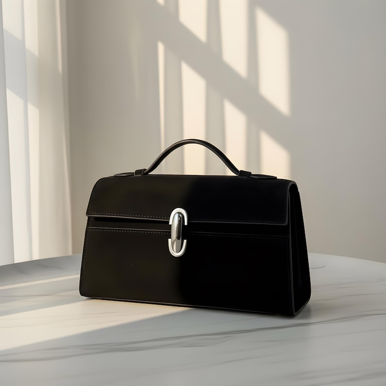 K&ouml;nnte beinhalten: Eine schwarze Handtasche mit einem oberen Griff und einem silberfarbenen Verschluss. Die rechteckige Tasche hat einen Klappenverschluss und steht auf einer wei&szlig;en Oberfl&auml;che. Die Tasche ist aus glattem Material gefertigt und hat ein strukturiertes Design.